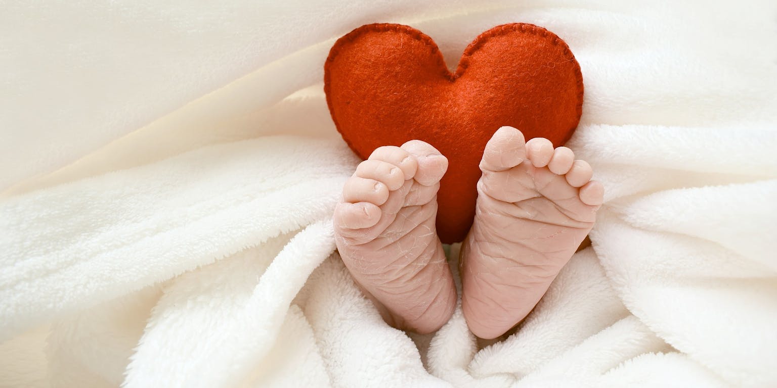 Een foto van een baby onder de dekens. Alleen de voetjes en een rood knuffelhart komen boven de dekens uit,