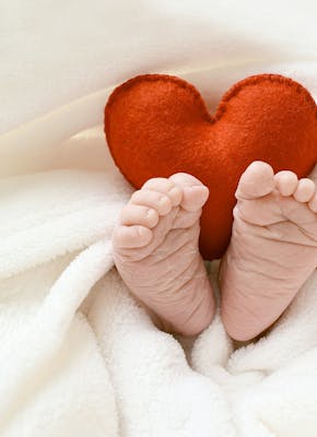 Een foto van een baby onder de dekens. Alleen de voetjes en een rood knuffelhart komen boven de dekens uit,