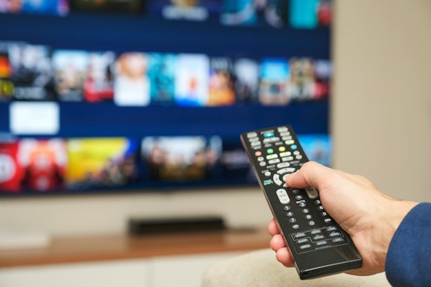 Een hand richt een afstandsbediening op een tv. Op het scherm is onscherp een streamingsdienst te zien, met plaatjes van verschillende films en series.