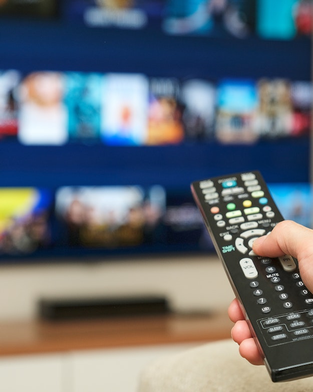 Een hand richt een afstandsbediening op een tv. Op het scherm is onscherp een streamingsdienst te zien, met plaatjes van verschillende films en series.