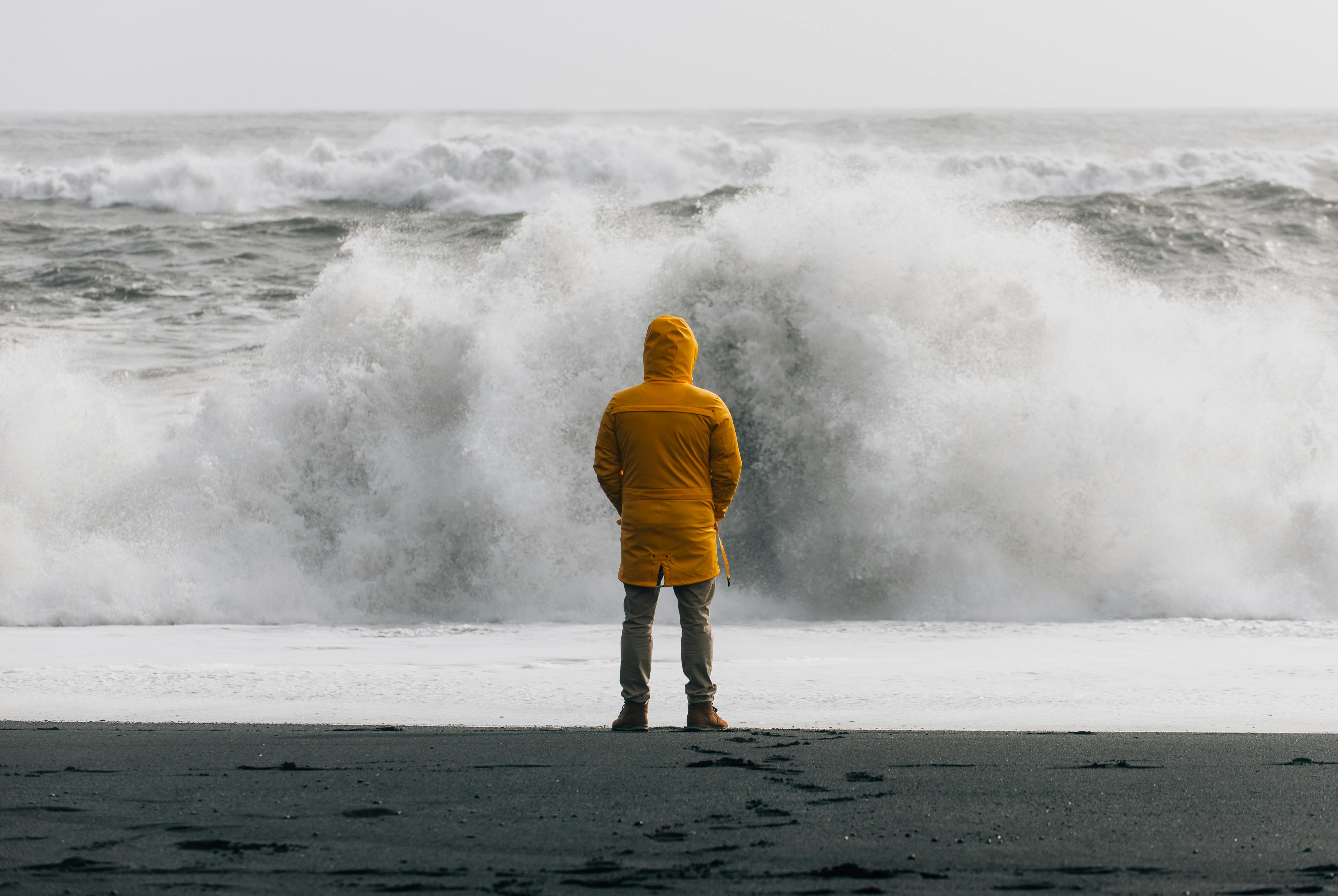 Een man met gele regenjas staat tijdens een storm aan zee en kijkt naar de hoog opspattende golven