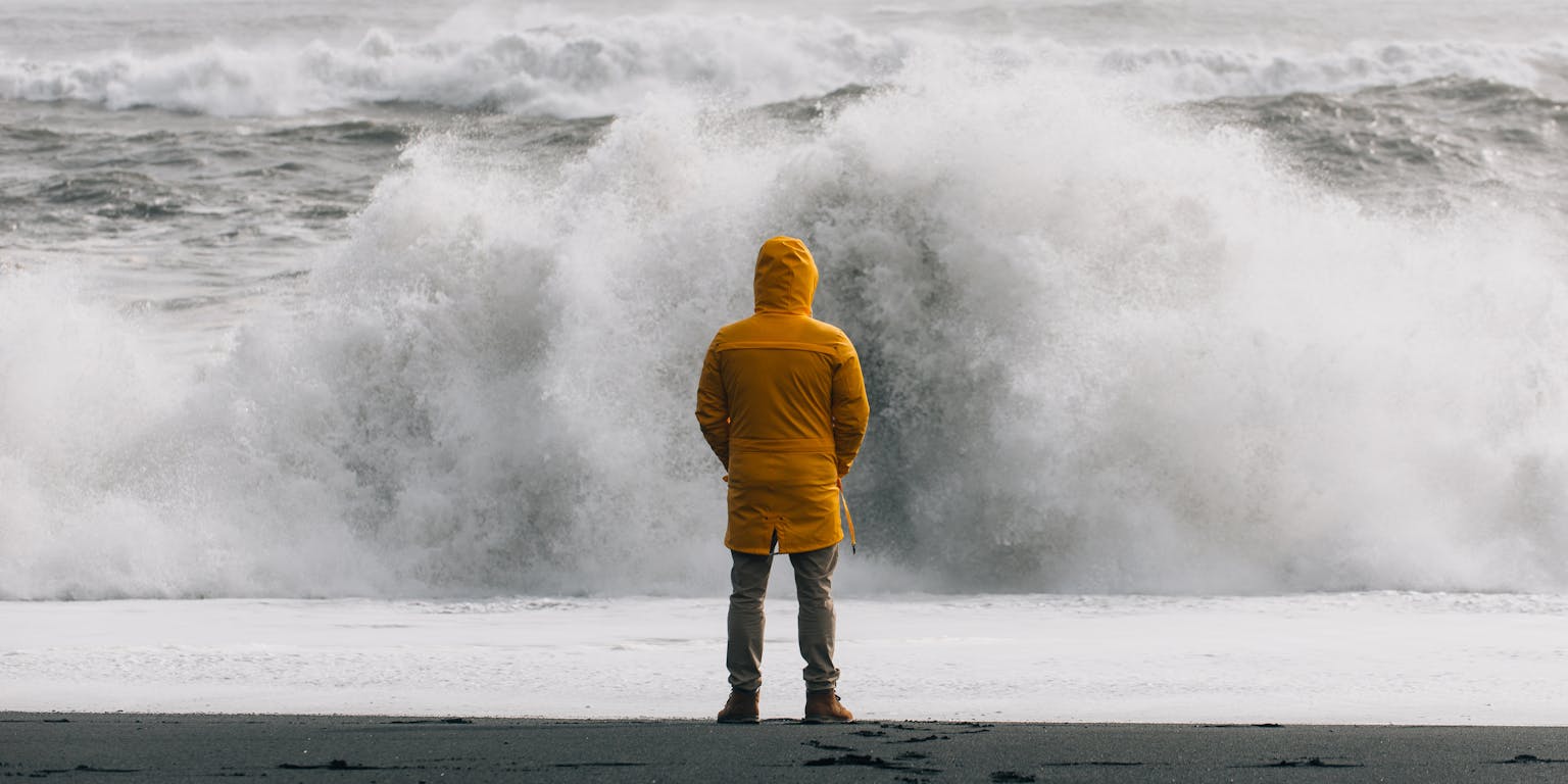 Een man met gele regenjas staat tijdens een storm aan zee en kijkt naar de hoog opspattende golven