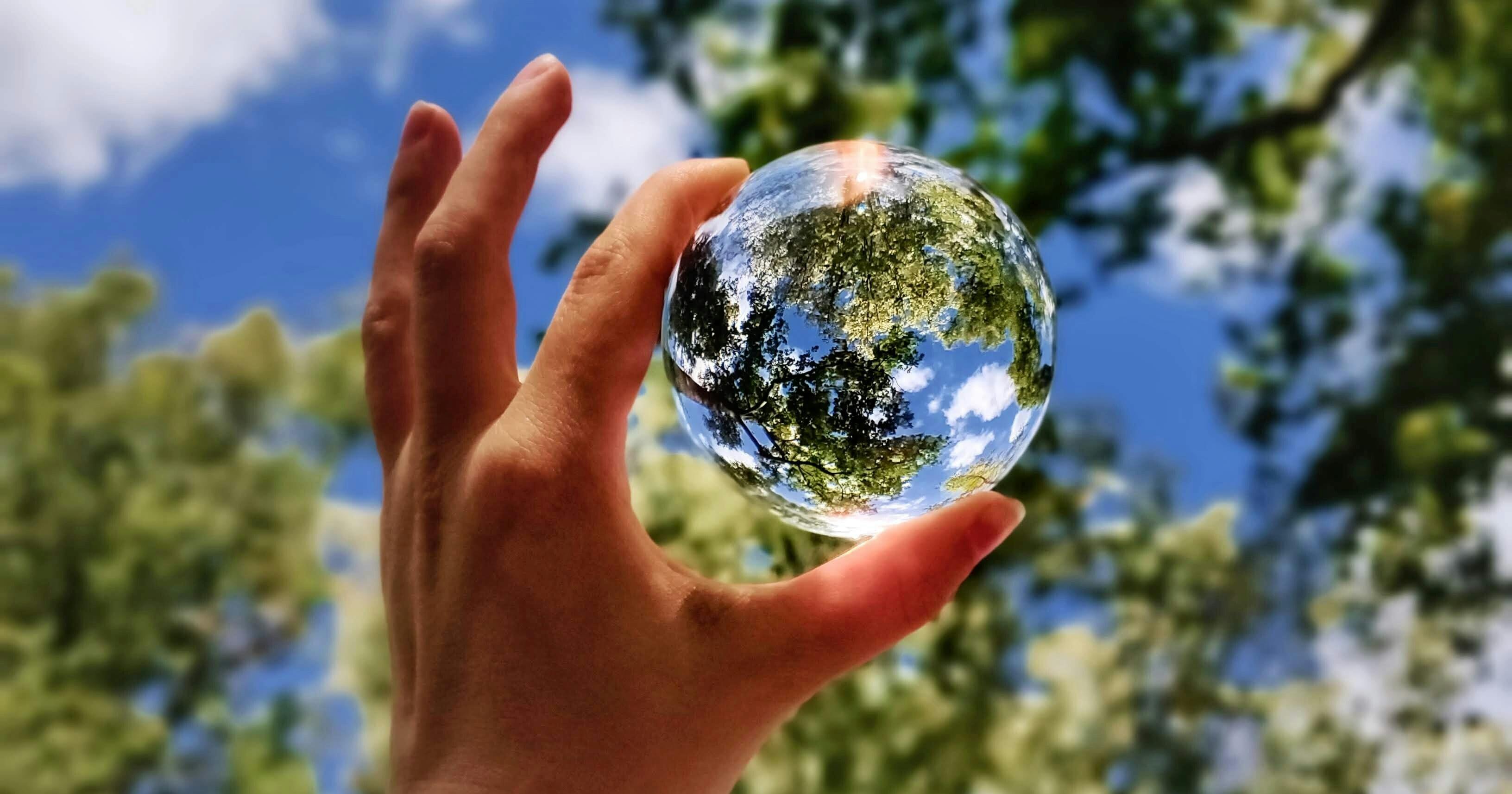 De hand van iemand die een glazen bol vasthoudt met daarin de weerspiegeling van een bladerdak