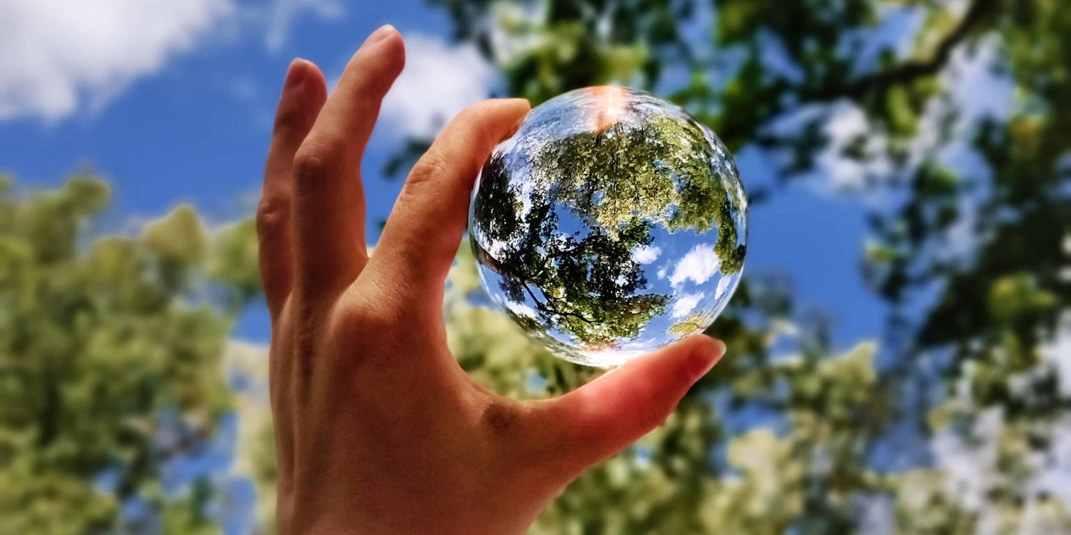 De hand van iemand die een glazen bol vasthoudt met daarin de weerspiegeling van een bladerdak