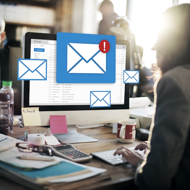 Een vrouw zit aan een rommelig bureau achter een computer. Op het scherm is een mailbox geopend. Enveloppen die ongelezen e-mails symboliseren, zweven voor het beeldscherm.