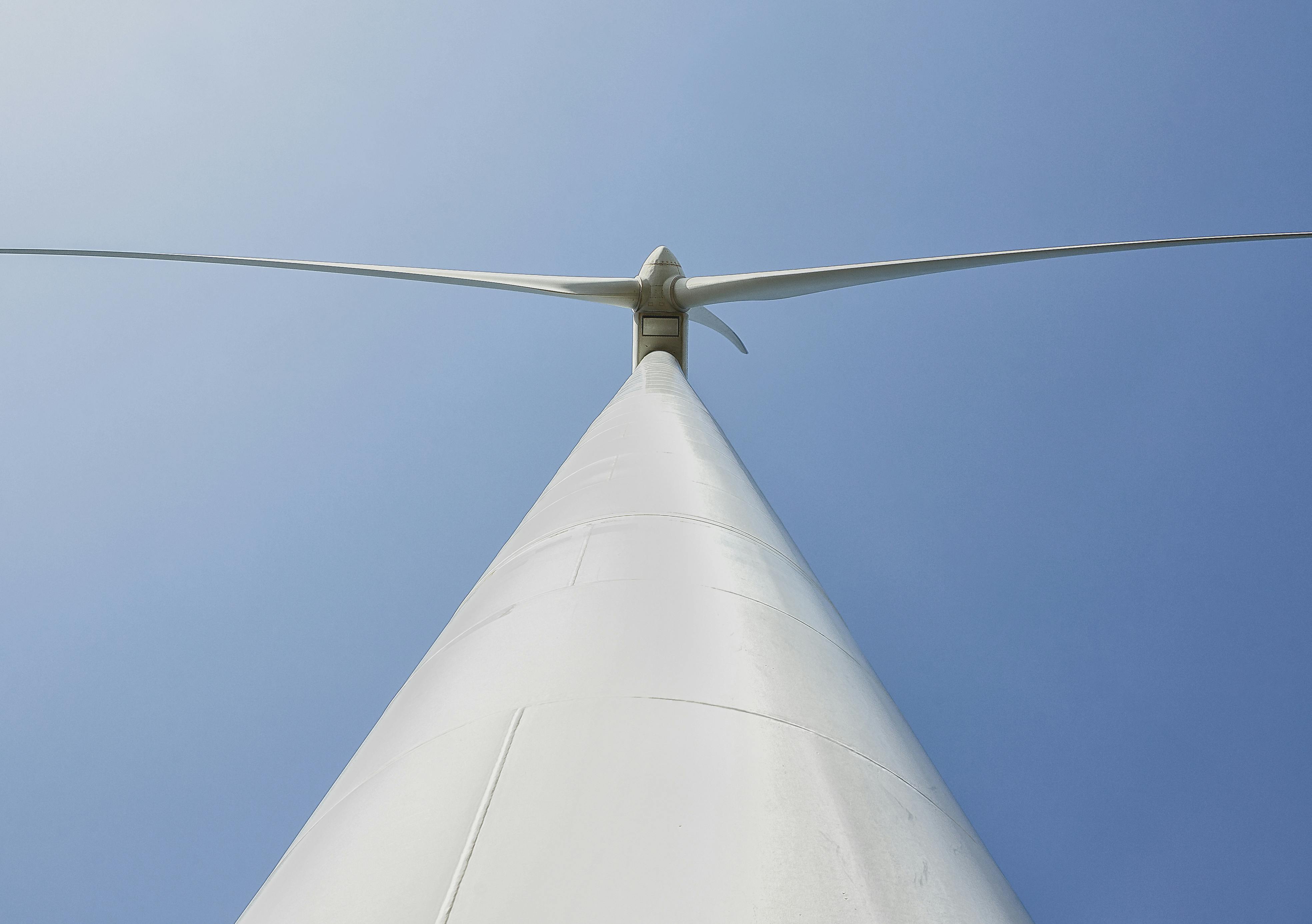 Een witte paal van een windmolen van onderaf gezien met twee turbinebladen die naar links en rechts uitsteken, tegen een blauwe lucht.
