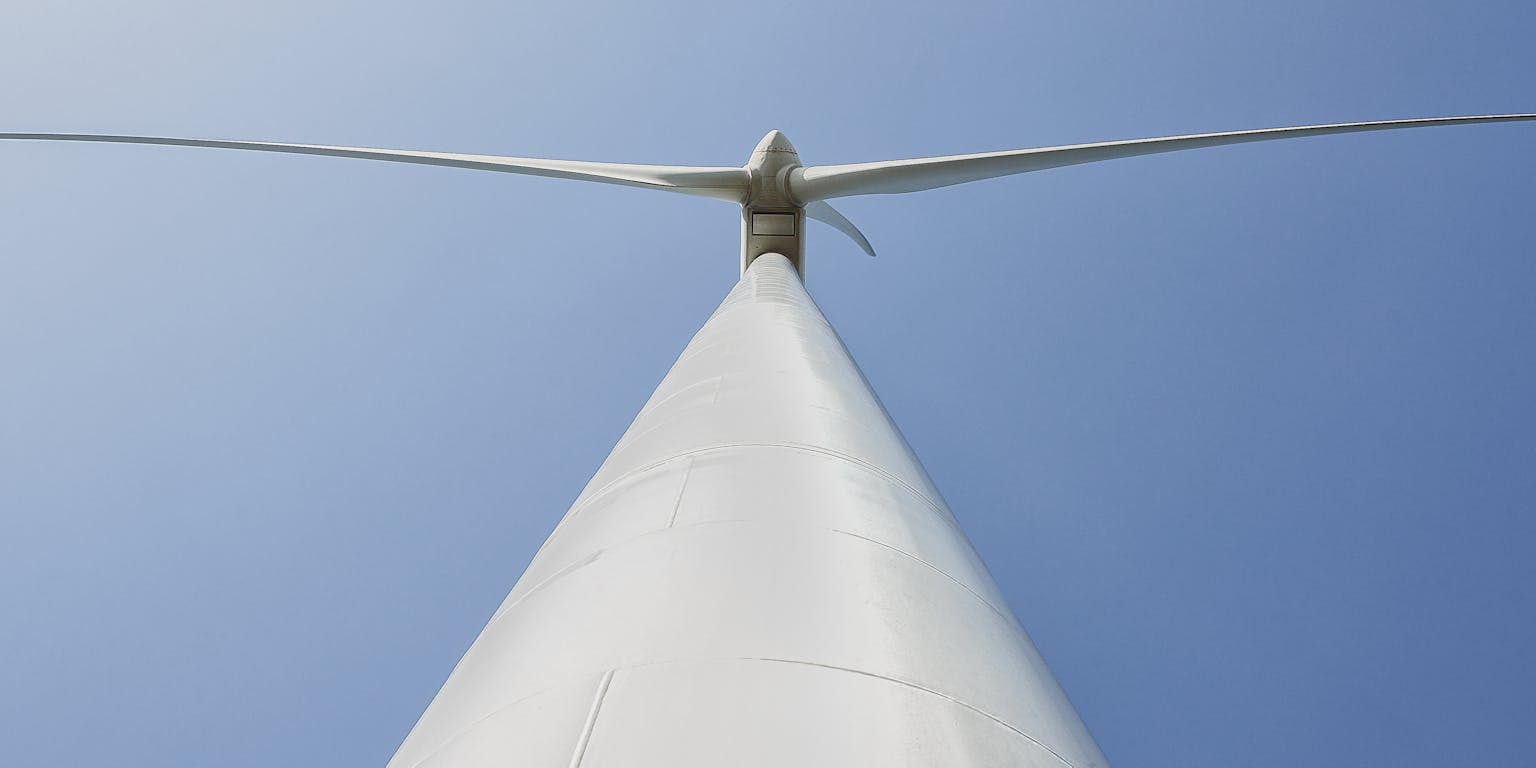 Een witte paal van een windmolen van onderaf gezien met twee turbinebladen die naar links en rechts uitsteken, tegen een blauwe lucht.