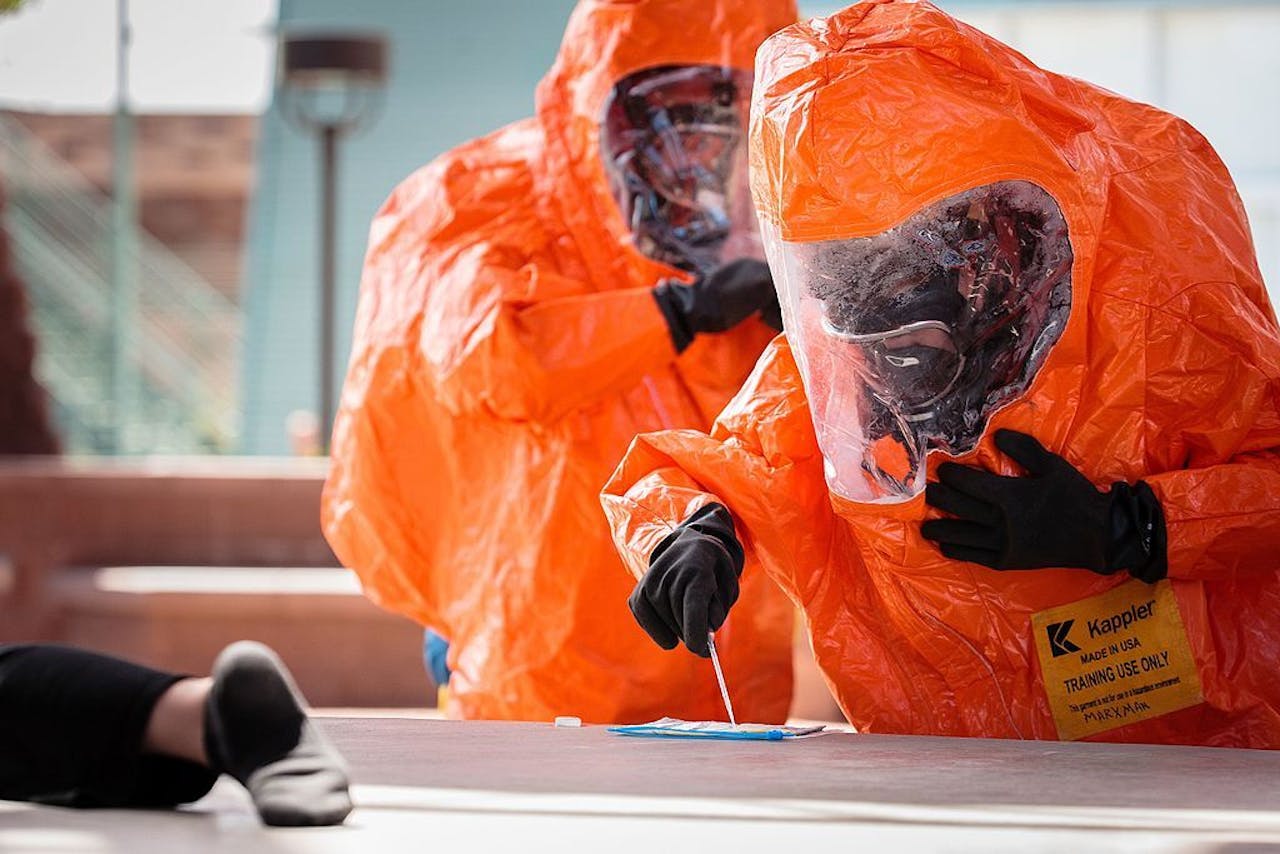Twee mensen in oranje beschermende kleding nemen een monster. Deze handeling vond plaats tijdens een oefening waarbij een aanval met chemische wapens werd nagebootst.