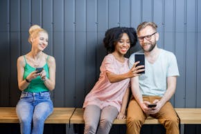 Een vrouw laat iets op haar smartphone zien aan een man, die ook een smartphone vasthoudt. Een andere vrouw met een smartphone in haar hand kijkt naar het tweetal. Ze lachen alle drie.