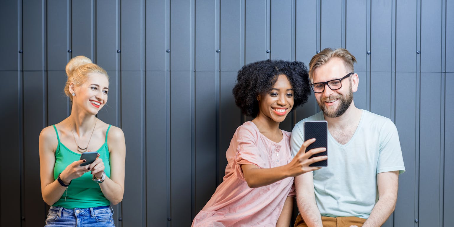 Een vrouw laat iets op haar smartphone zien aan een man, die ook een smartphone vasthoudt. Een andere vrouw met een smartphone in haar hand kijkt naar het tweetal. Ze lachen alle drie.