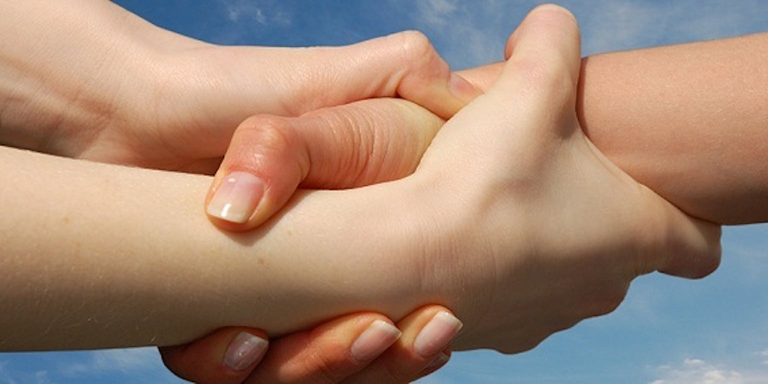 Een close-up van twee mensen die met hun hand elkaars pols vasthouden. Op de achtergrond is een blauwe lucht te zien.