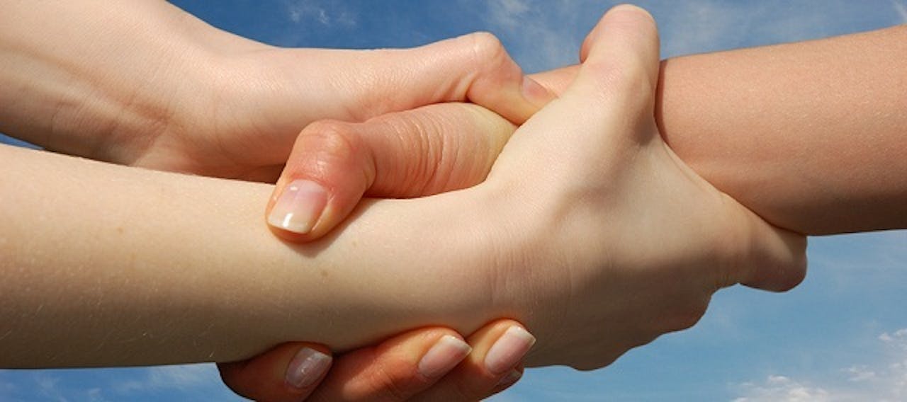 Een close-up van twee mensen die met hun hand elkaars pols vasthouden. Op de achtergrond is een blauwe lucht te zien.