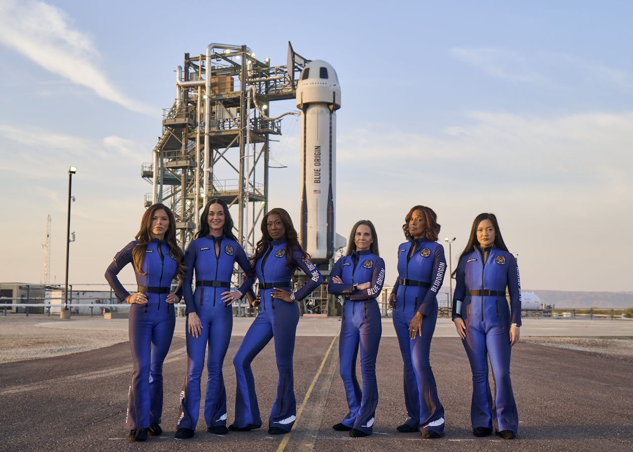 Zes vrouwen in een blauw pak op asfalt met op de achtergrond een raket op een lanceerplatform.
