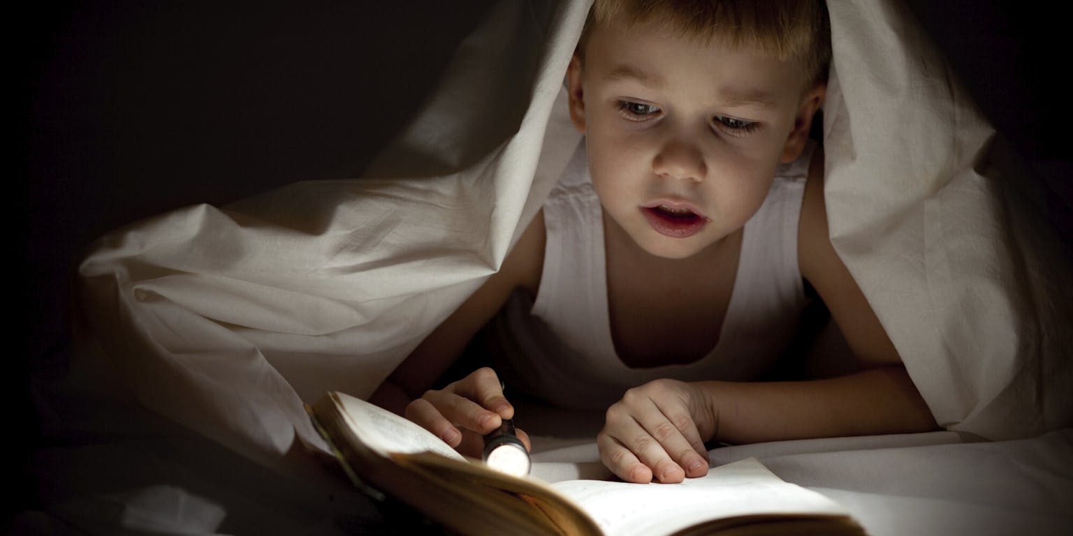 Een jongen leest een boek onder een deken met een zaklamp.