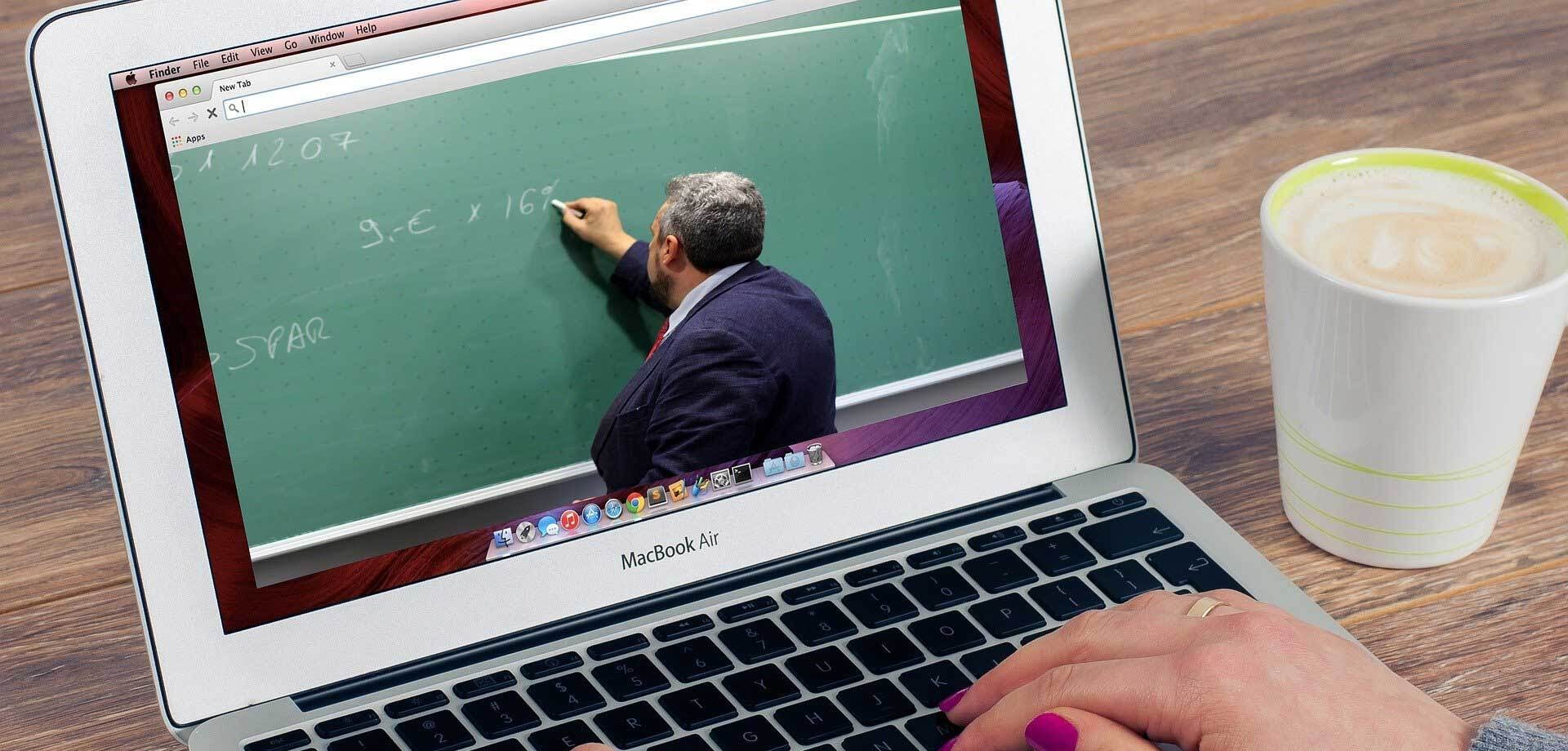 Een persoon die een laptop gebruikt om online een les van school te volgen. Op het beeldscherm is te zien dat er een persoon bij een schoolbord staat.