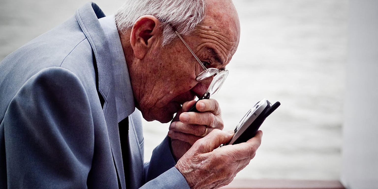 Een oudere man kijkt naar zijn mobiele telefoon met een vergrootglas in zijn hand.