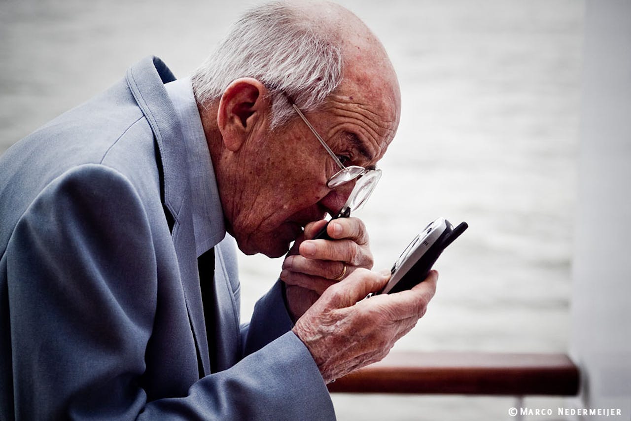 Een oudere man kijkt naar zijn mobiele telefoon met een vergrootglas in zijn hand.