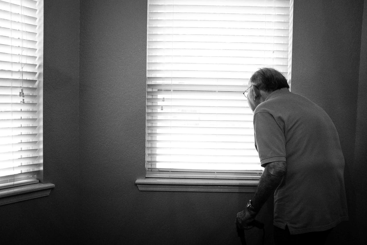 Een oudere man tuurt door een raam waar lamellen voor hangen.