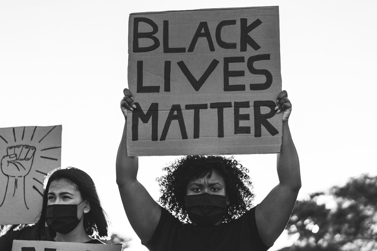 Een donkere vrouw met zwart mondkapje houdt een kartonnen bord met de tekst 'Black Lives Matter' omhoog. Naast haar staat een vrouw, eveneens met een zwart mondkapje. Een bord met een getekende vuist is zichtbaar.