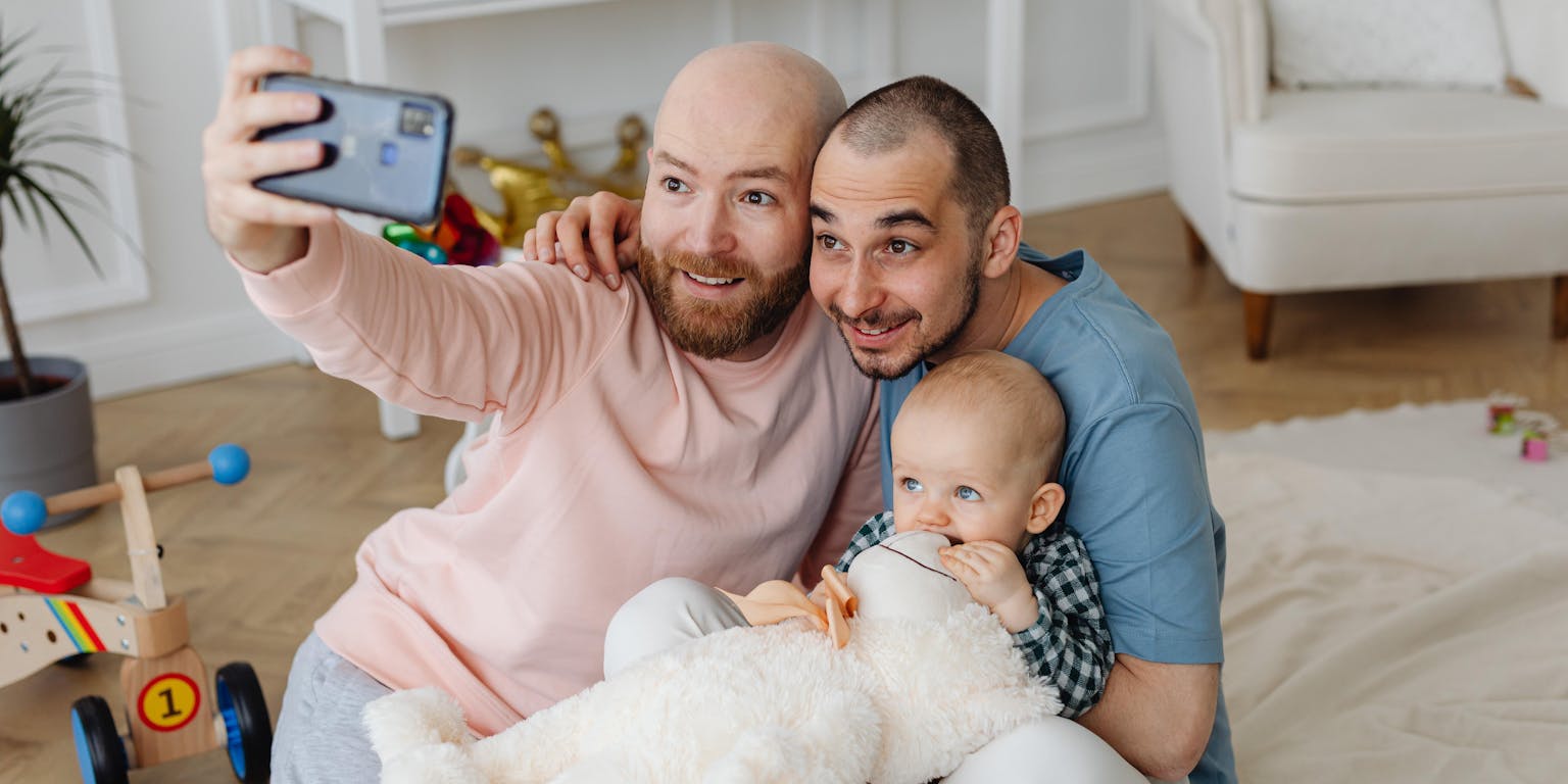 Twee mannen en een baby maken een selfie in een woonkamer.