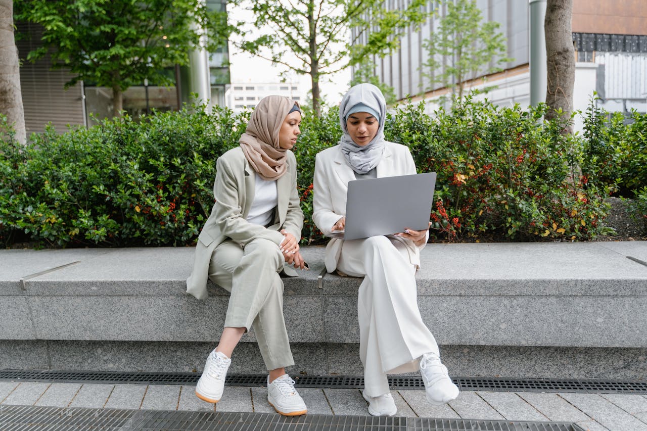 Twee vrouwen die een hoofddoek dragen, zitten op een bankje en gebruiken een laptop.