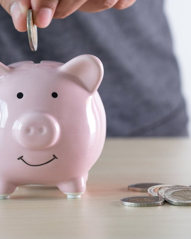 Een hand stopt een munt in een roze spaarvarken. Naast het varken liggen nog meer munten. Een hand stopt een munt in een roze spaarvarken. Naast het varken liggen nog meer munten.