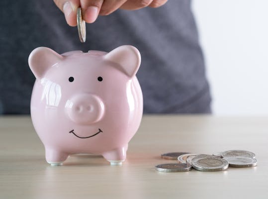 Een hand stopt een munt in een roze spaarvarken. Naast het varken liggen nog meer munten.