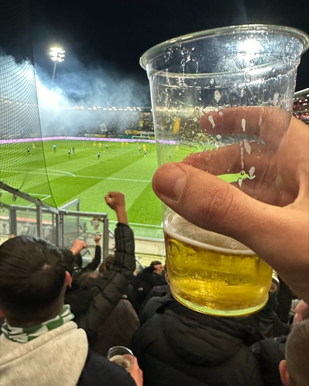 Een hand houdt een plastic beker met bier vast. Op de achtergrond is een tribune met supporters en een voetbalveld te zien. Een hand houdt een plastic beker met bier vast. Op de achtergrond is een tribune met supporters en een voetbalveld te zien.