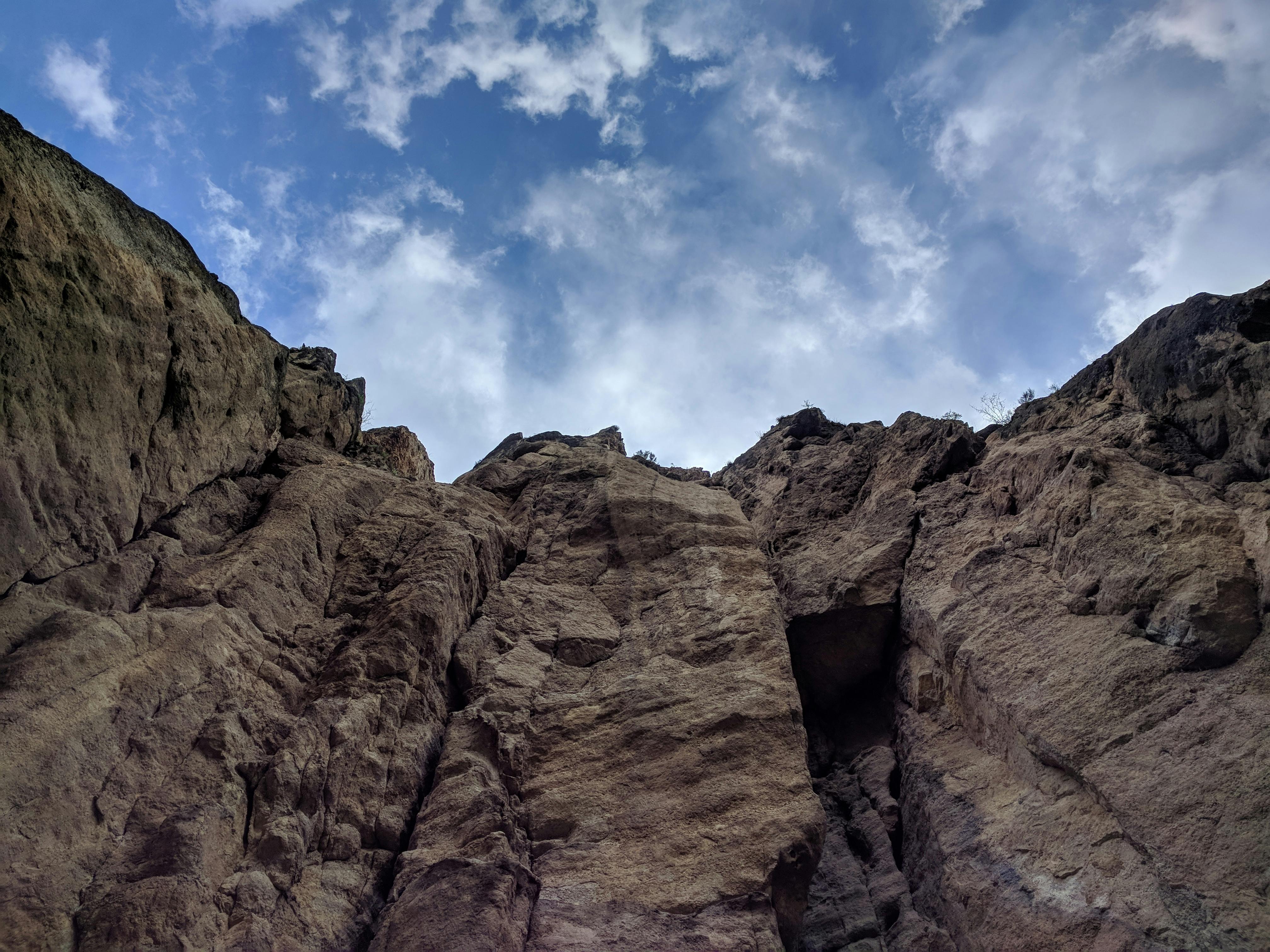 Bruin-grijze rots vanaf onder gezien tegen blauwe lucht met witte wolkjes.