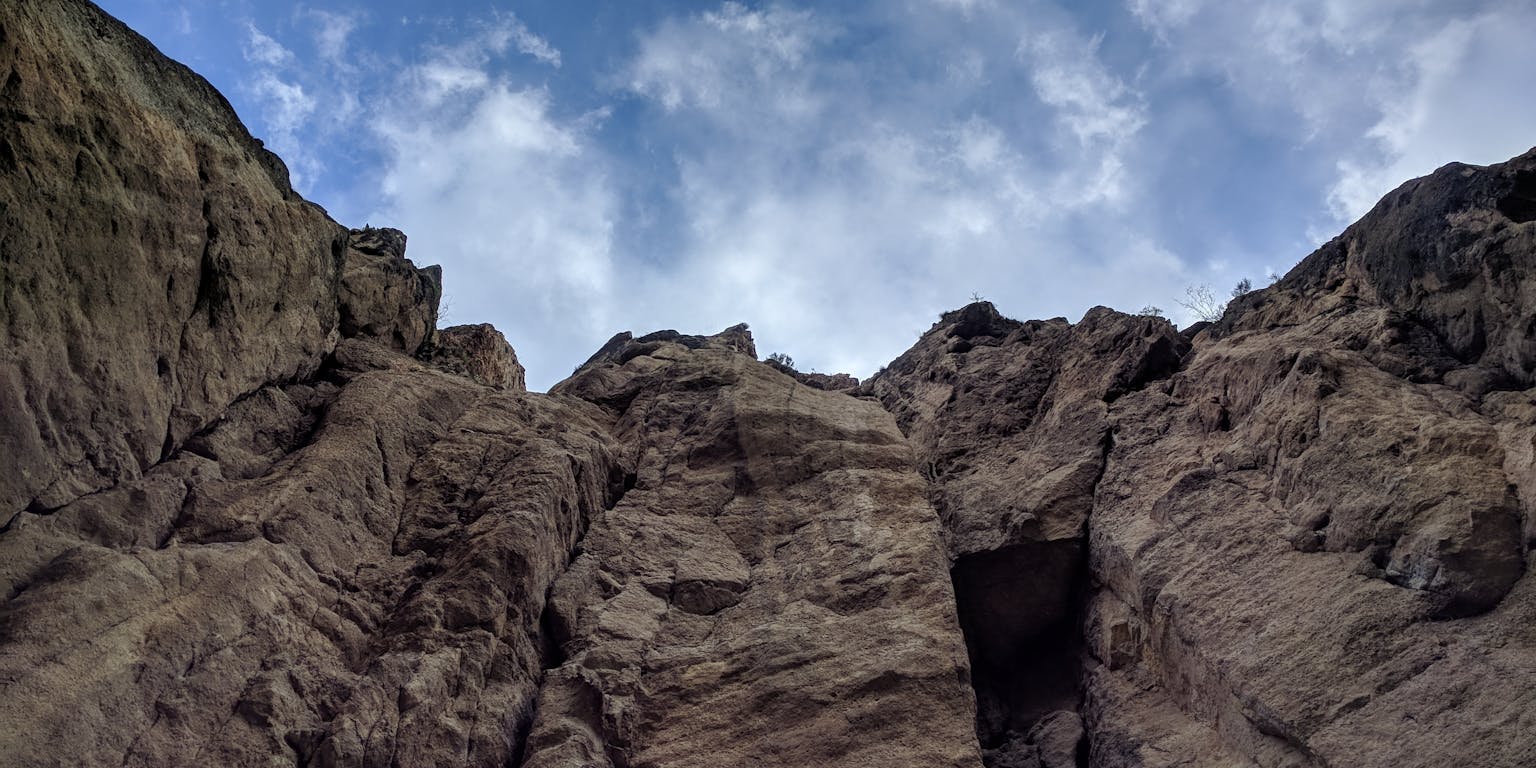 Bruin-grijze rots vanaf onder gezien tegen blauwe lucht met witte wolkjes.