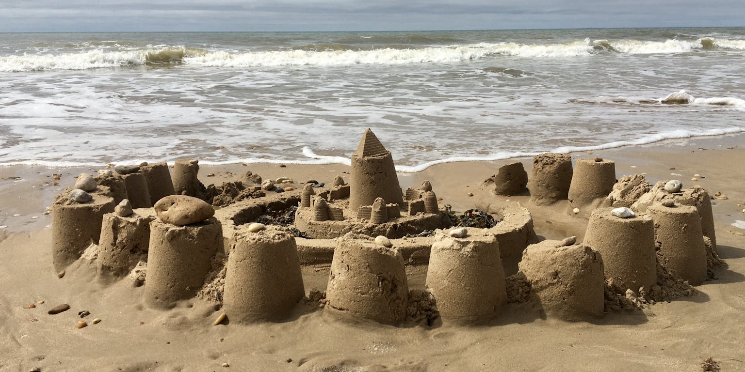 Half in elkaar gezakt zandkasteel met op de achtergrond wat zeegolven