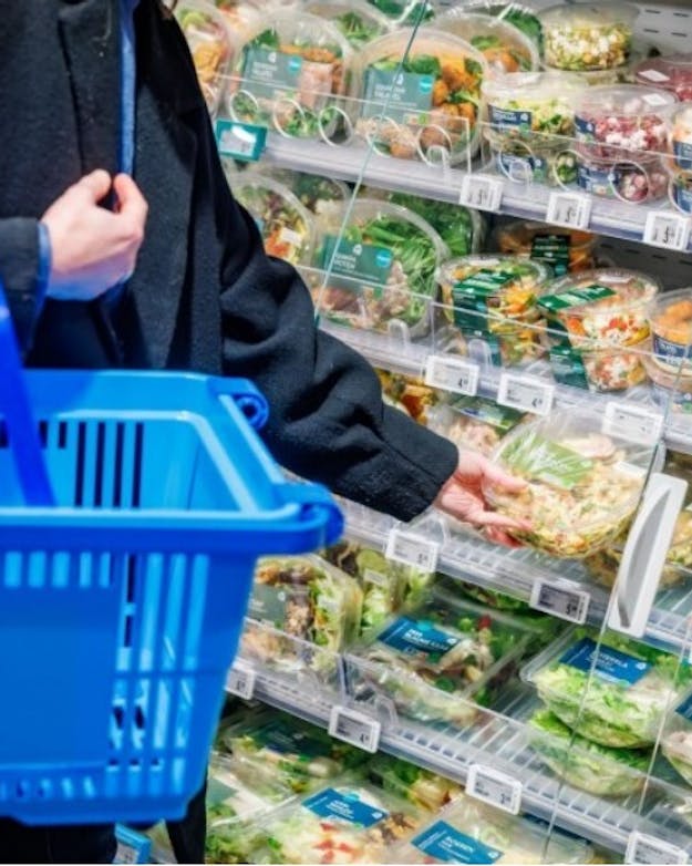 Een klant in een supermarkt heeft een blauw winkelmandje met daarop het logo van Albert Heijn aan de arm hangen. De klant pakt een voorverpakte salade uit een koelvak. Een klant in een supermarkt heeft een blauw winkelmandje met daarop het logo van Albert Heijn aan de arm hangen. De klant pakt een voorverpakte salade uit een koelvak.