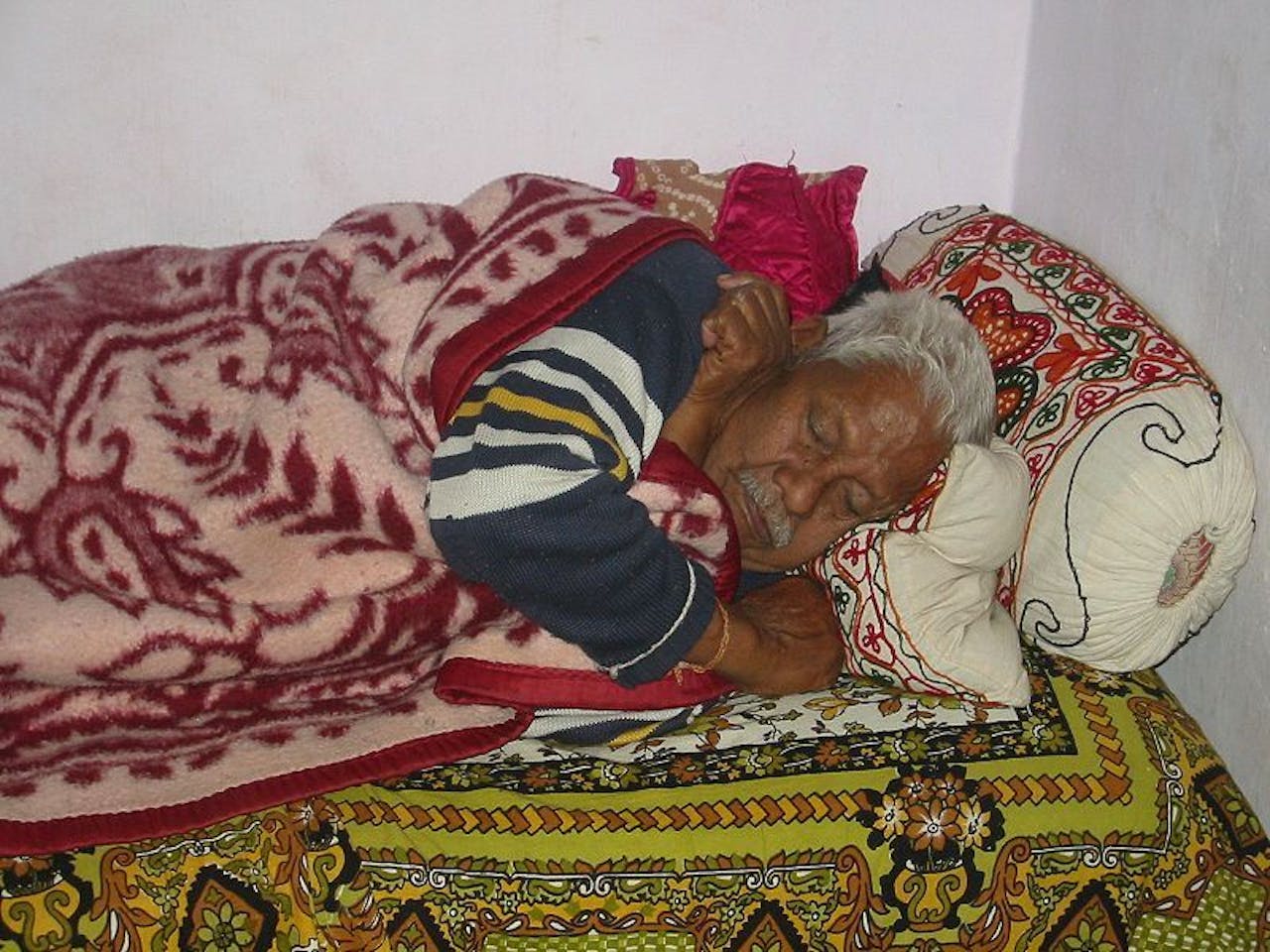 Een oude man slaapt op een bed onder een gekleurde deken.