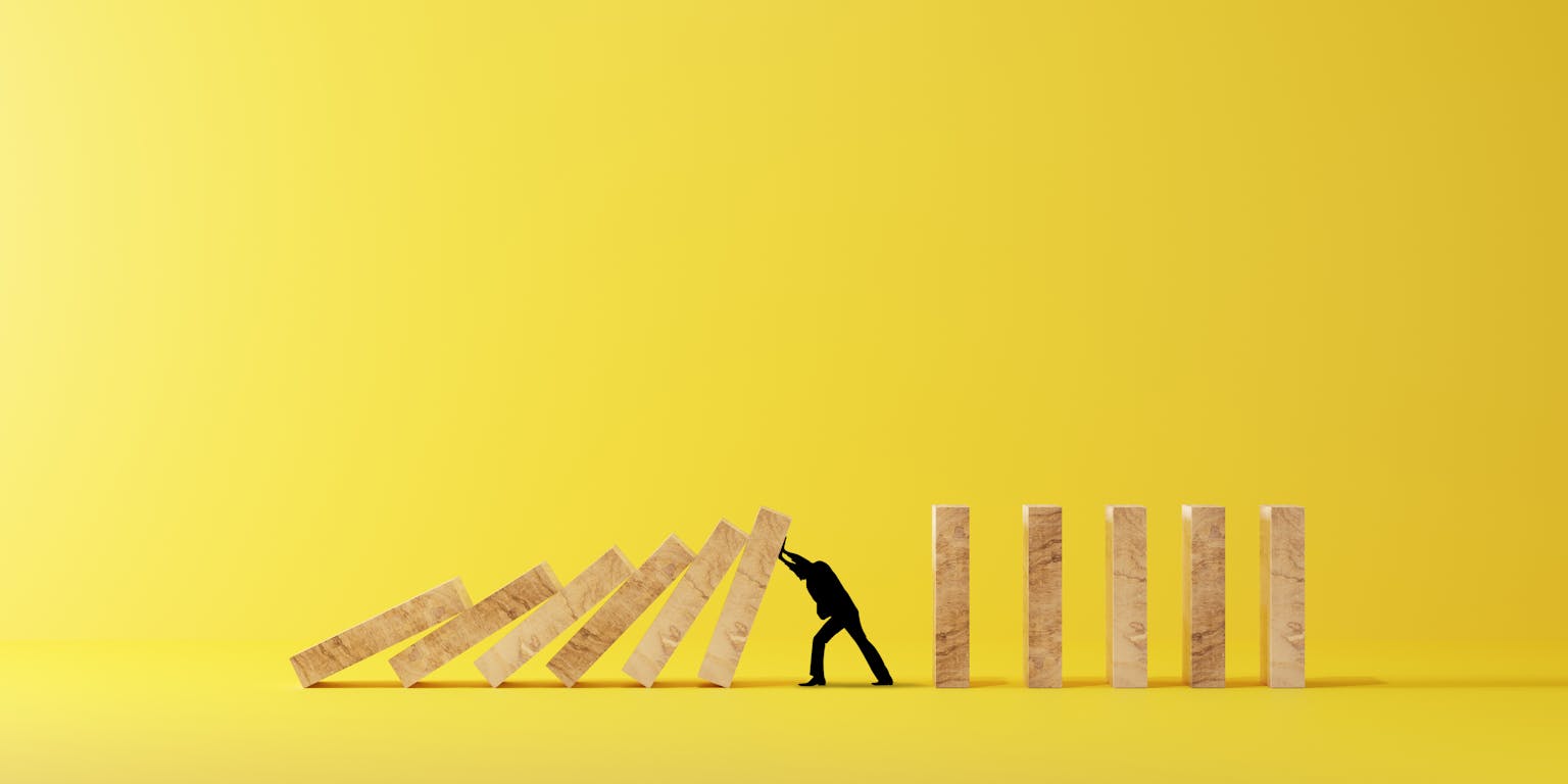 Een miniatuur silhouet van een man probeert een rij houten dominostenen tegen te houden, maar wordt er toch door bedolven