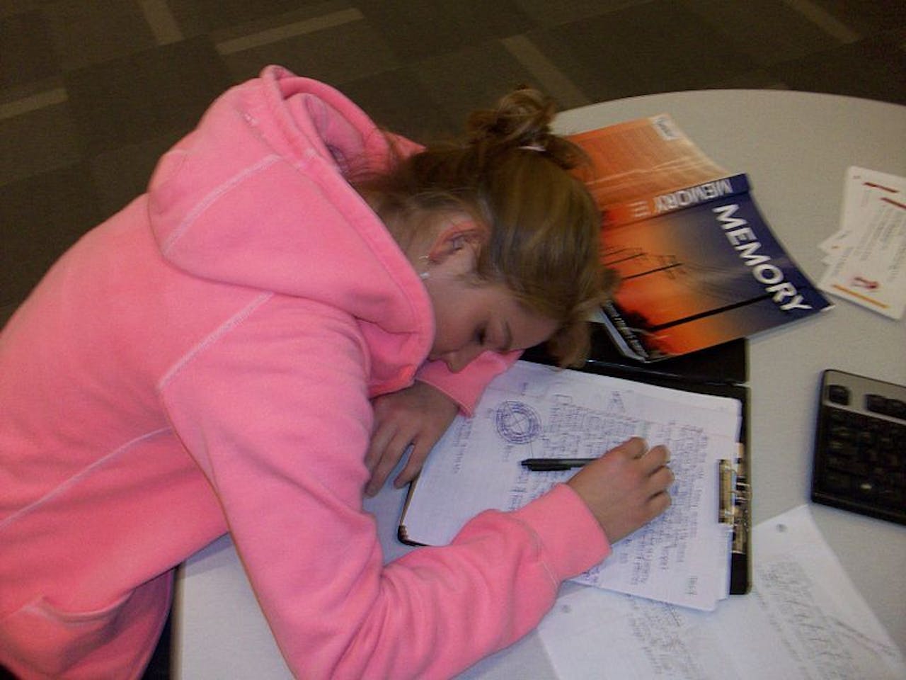 Een student zit aan tafel met een boek en aantekeningen. De student slaapt met haar hoofd op tafel.