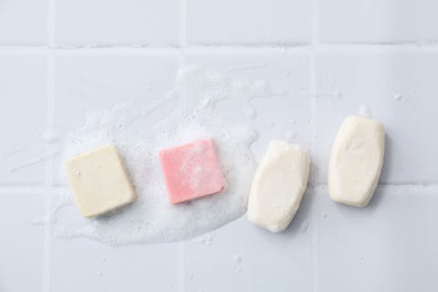 Shampooflessen zijn een toepassing van plastic die niet per se nodig is. Ouderwetse zeepblokken volstaan ook. Vier zeepblokken (drie witte en één roze) liggen met schuim op witte tegeltjes.