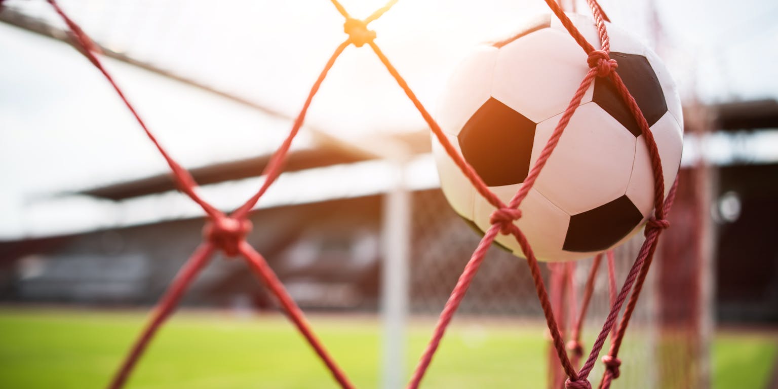 Voetbal belandt in het rode net van een goal. Op de achtergrond is wazig de grasmat en de tribune van het stadion te zien.