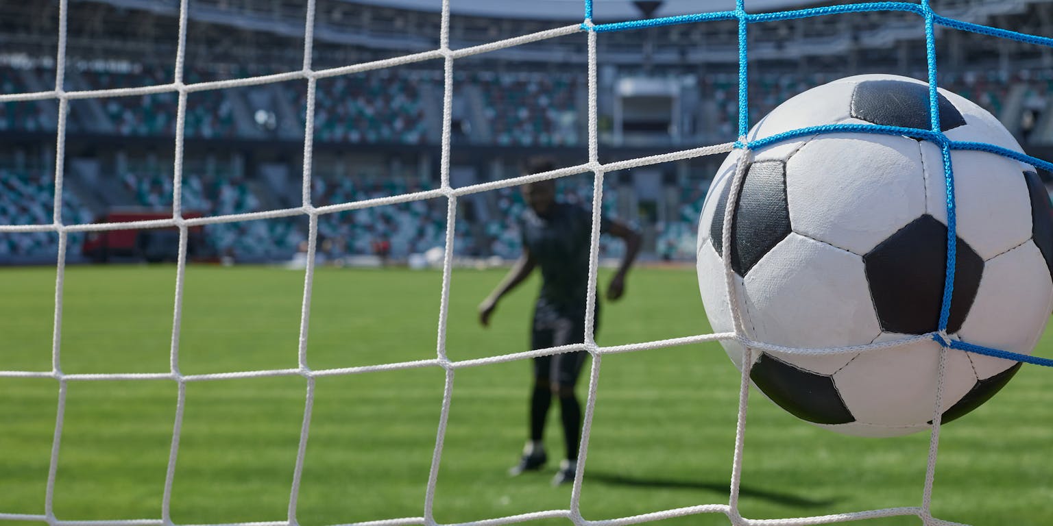 Voetbal in het net van een goal. Door het net zie je een voetbalspeler staan in een stadion met supporters op de tribunes.