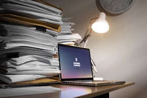 Stilleven met twee flinke stapels papier en documenten en een laptop op een bureau. Een witte bureaulamp verlicht de laptop. Aan de muur hangt een klok.