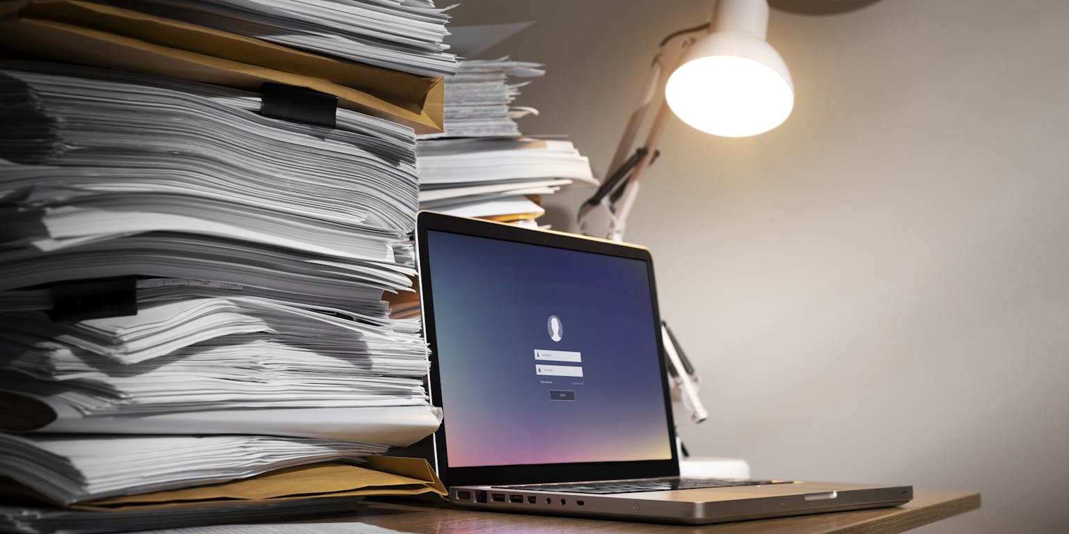 Stilleven met twee flinke stapels papier en documenten en een laptop op een bureau. Een witte bureaulamp verlicht de laptop. Aan de muur hangt een klok.