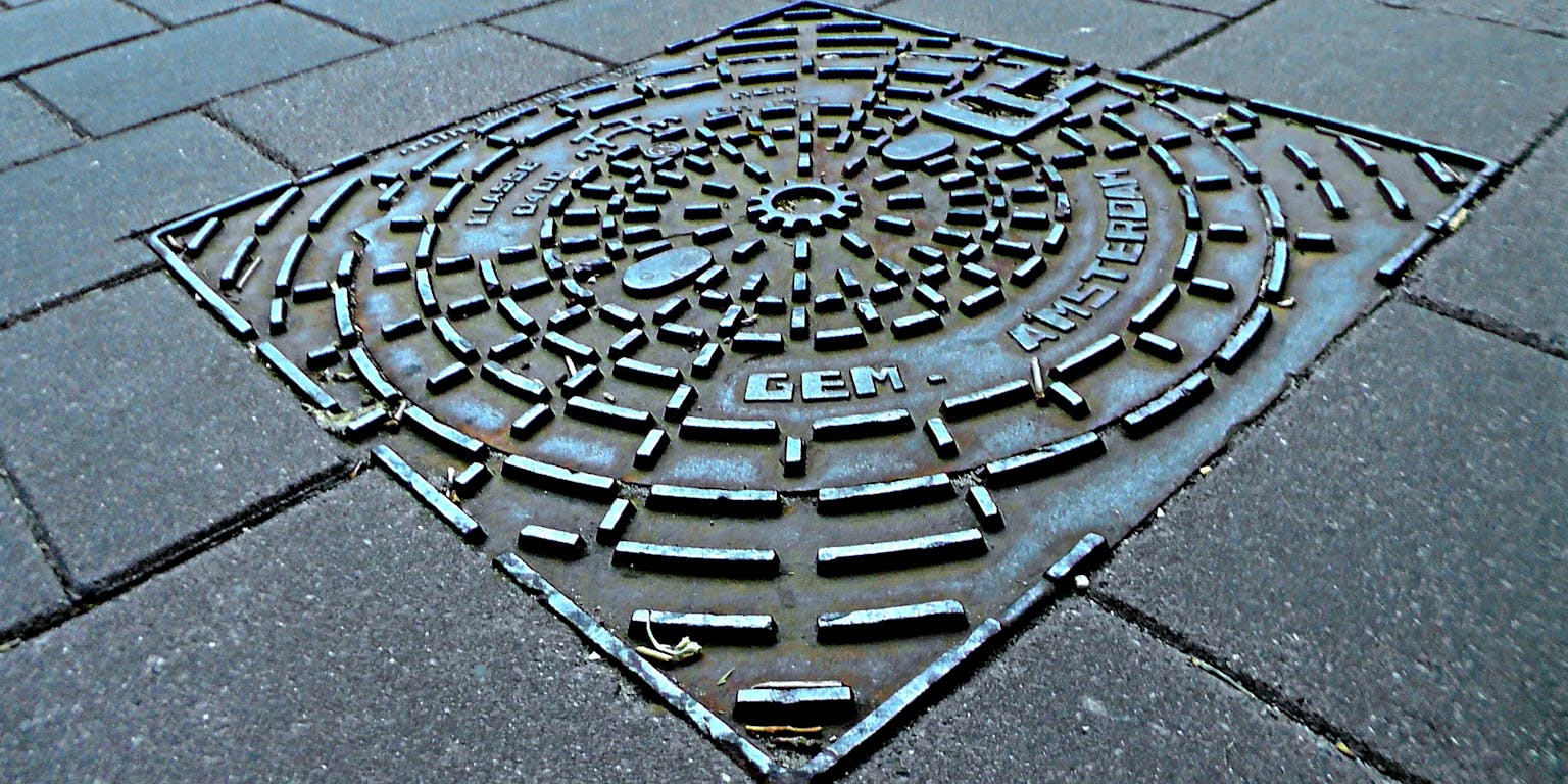 Een putdeksel van metaal met daarop de tekst 'Gem. Amsterdam' ligt tussen stoeptegels.