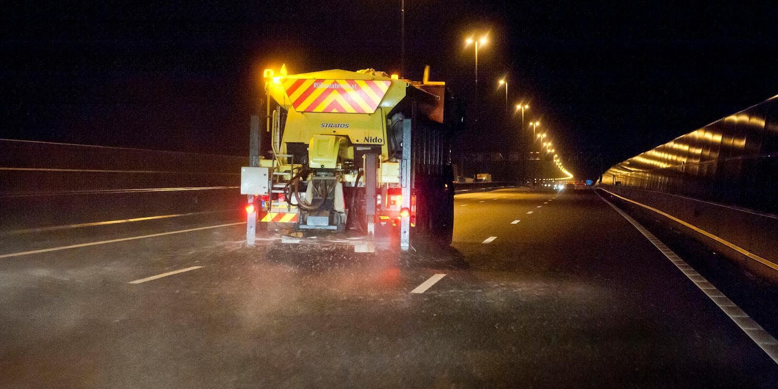 Een gele vrachtwagen van achter gezien op een lege snelweg in het donker strooit zout. De lantaarnpalen langs de weg branden.