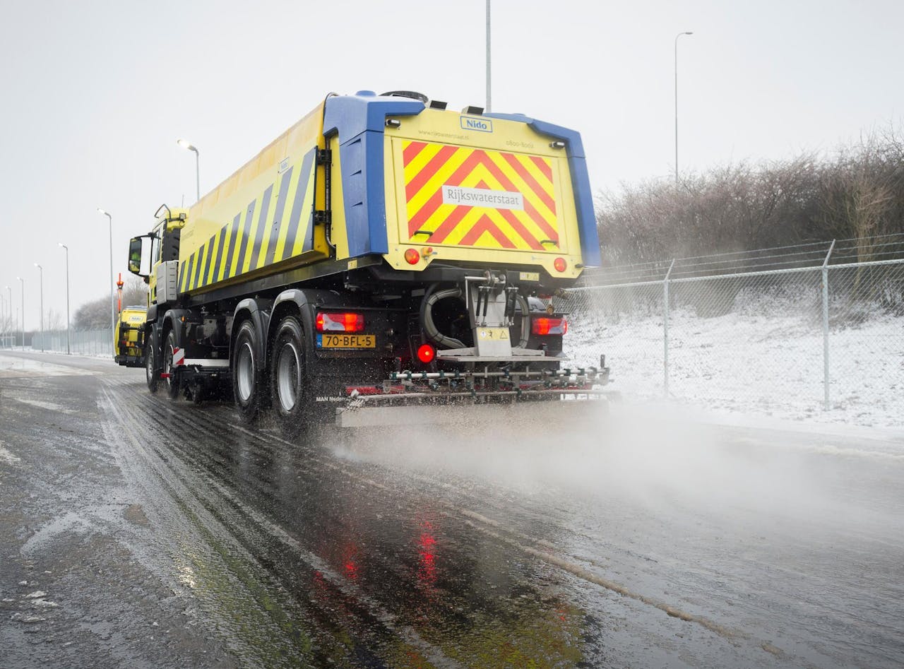 Een gele vrachtwagen rijdt in winters weer over de weg en strooit zout. In de berm ligt sneeuw.