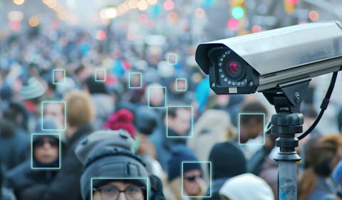 Computers zijn goed in het detecteren van groepen mensen die dichtbij elkaar staan. Een wazige menigte op straat met op de voorgrond een surveillancecamera. Rond verschillende gezichten is een vierkant getekend.