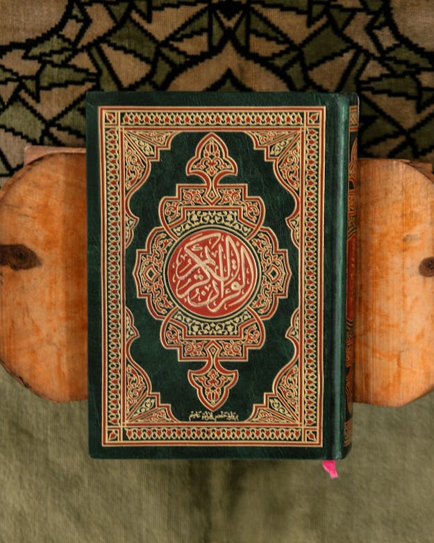 Een religieus boek, zoals de Koran, staat vol voorschriften en doordachte ideeën over hoe de goddelijke wereld in elkaar zit. Hierdoor is er minder ruimte voor genderdiversiteit in de verhalen. Goud met groene Koran op houten standaard op groenig tapijt.