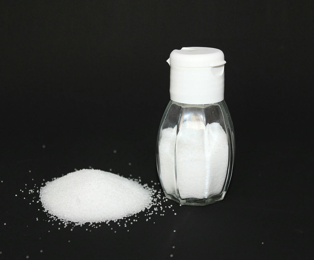 Een potje tafelzout, gevuld met zout. Er ligt ook zout los voor het potje op een zwarte achtergrond.