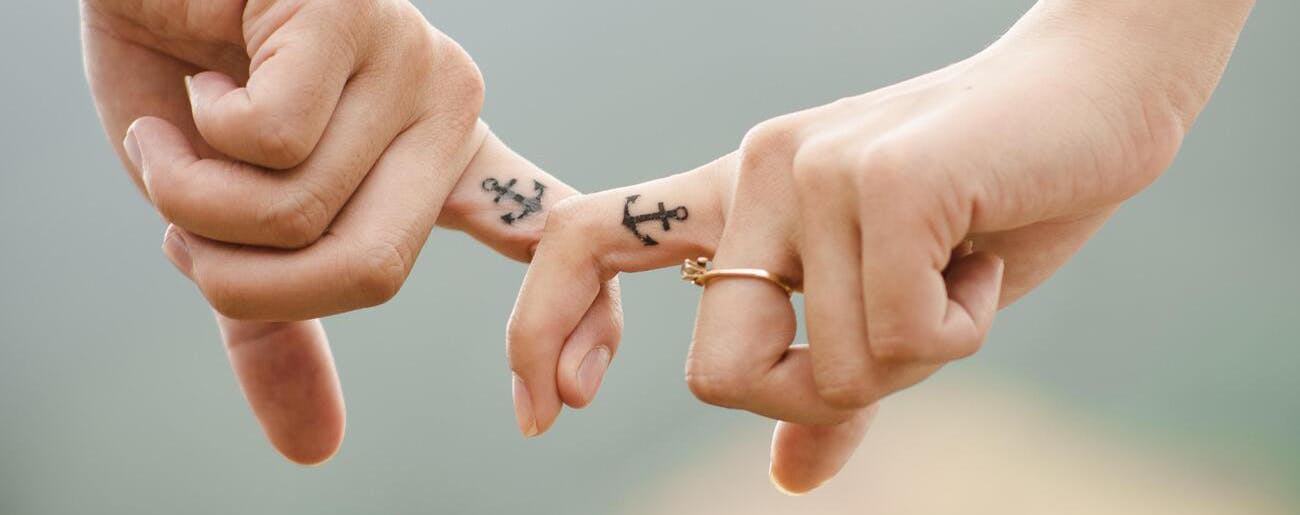 Twee mensen haken hun vinger met daarop een tatoeage van een anker in elkaar.