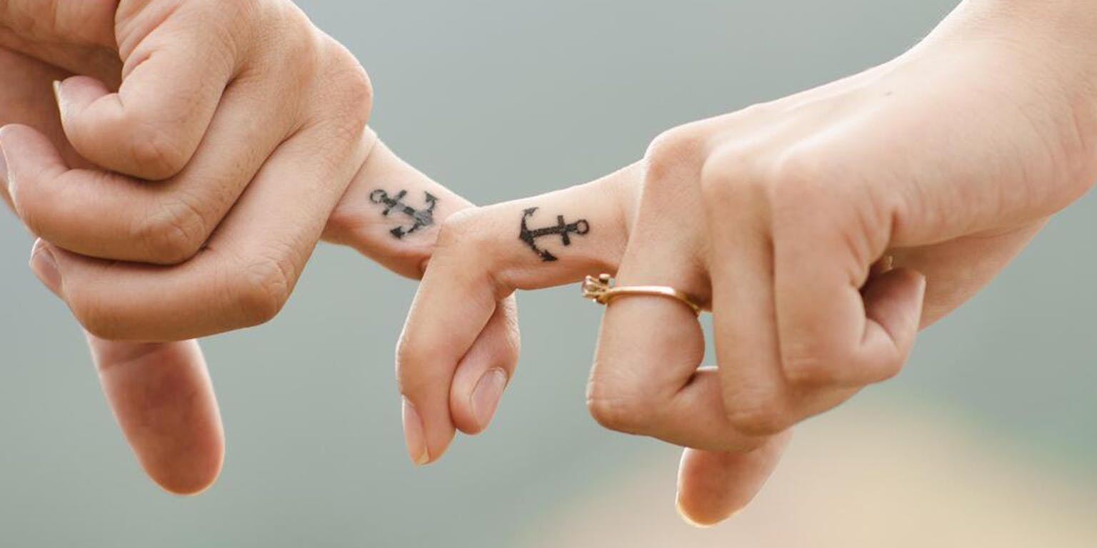 Twee mensen haken hun vinger met daarop een tatoeage van een anker in elkaar.