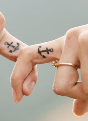 Twee mensen haken hun vinger met daarop een tatoeage van een anker in elkaar.