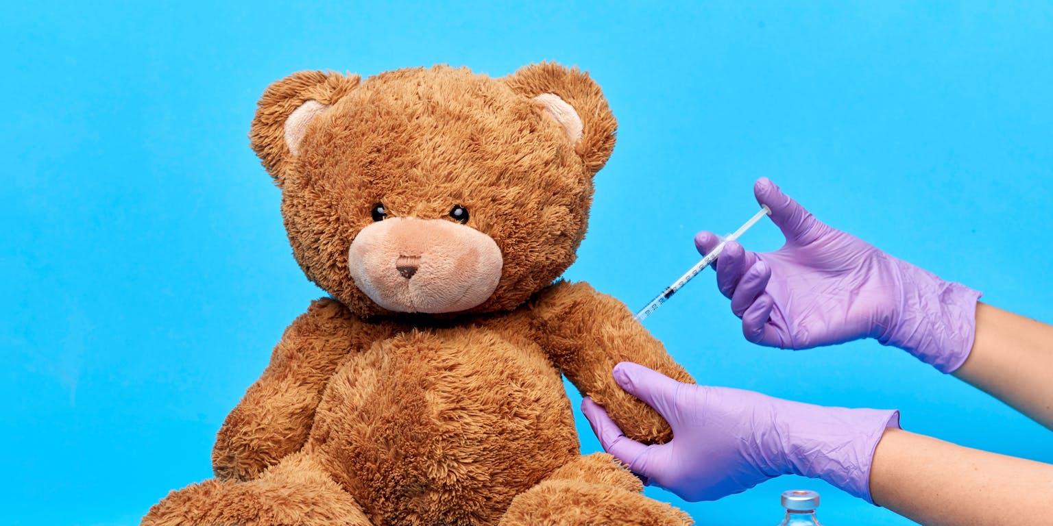 Een teddybeer krijgt een spuitje van iemand met handschoenen aan en witte doktersjas aan